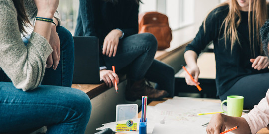 Brainstorming Vier Menschen sitzen an einem Tisch, die Gesichter sind nicht im Bild. Sie halten Stifte in der Hand und sehen nachdenkend aus. Eine Hand schreibt etwas auf das Plakat auf dem Tisch, welches sehr unstrukturiert gestaltet ist. Auf dem Tisch sind noch mehr Stifte und eine Tasse. Auf der Sitzbank erkennt man einen geöffneten Laptop und einen Rucksack im Hintergrund.